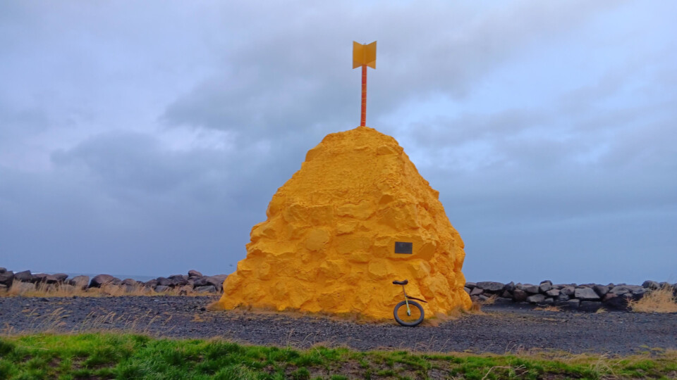 A large yellow mound of rock and concrete, with a yellow metal "flag" on top (Suðurnesvarða, "the cairn/beacon of Suðurnes", which historically served as a navigational waymarker). Leaning against it is a unicycle on black dirt, with grass in the foreground. The sky is gray and cloudy with small bits of light blue showing through.