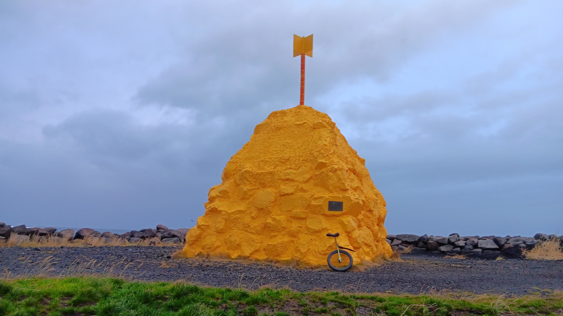 A large yellow mound of rock and concrete, with a yellow metal "flag" on top (Suðurnesvarða, "the cairn/beacon of Suðurnes" and surved as a navigational waymarker). Leaning against it is a unicycle on black dirt, with grass in the foreground. The sky is gray and cloudy with small bits of light blue showing through.