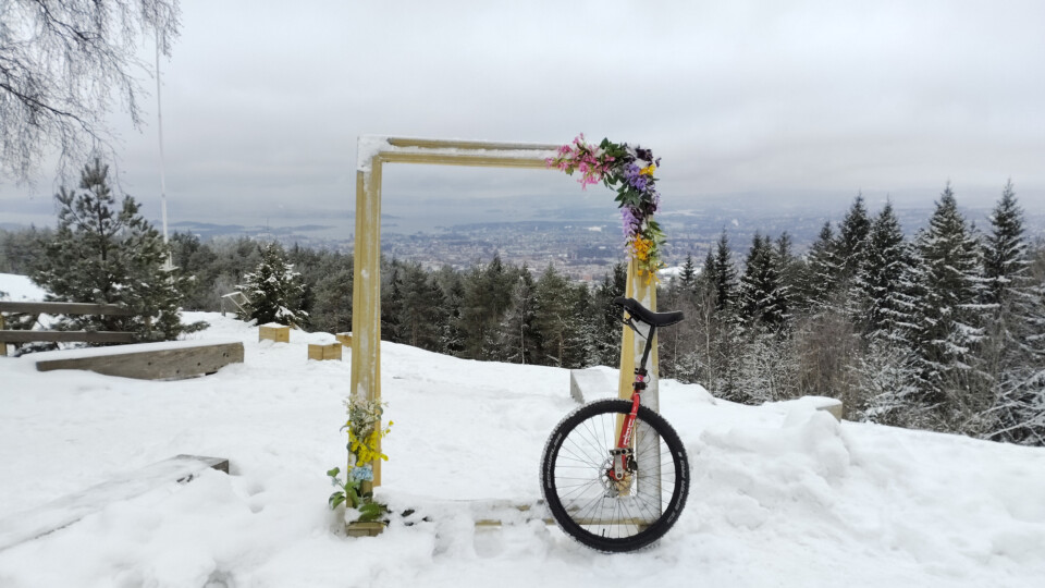 A unicycle leaning against a picture frame. There is snow on the ground and a view out over Oslo