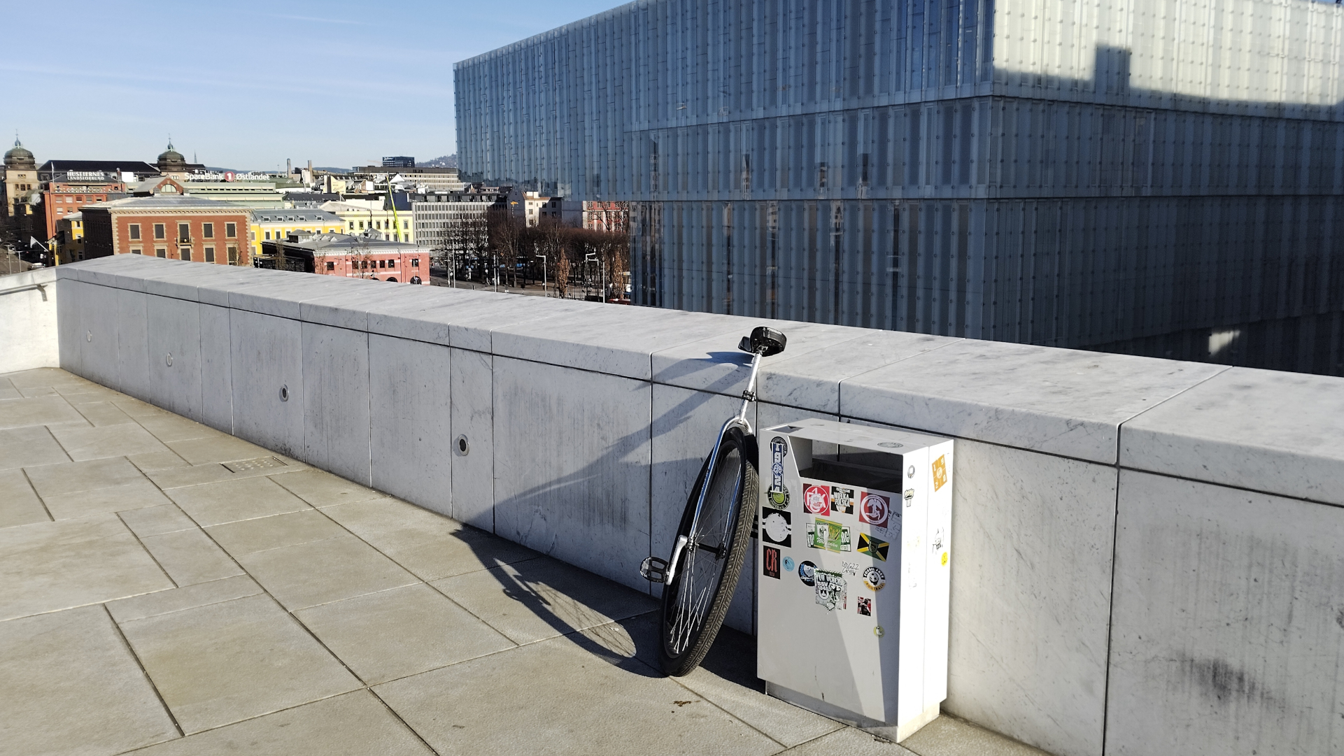 Large wheeled unicycle in front of a white stone wall next to a rectangular trash can covered in stickers. A big glass building is just past that in the near foregorund and beyong that other city buildings. Blue skies.