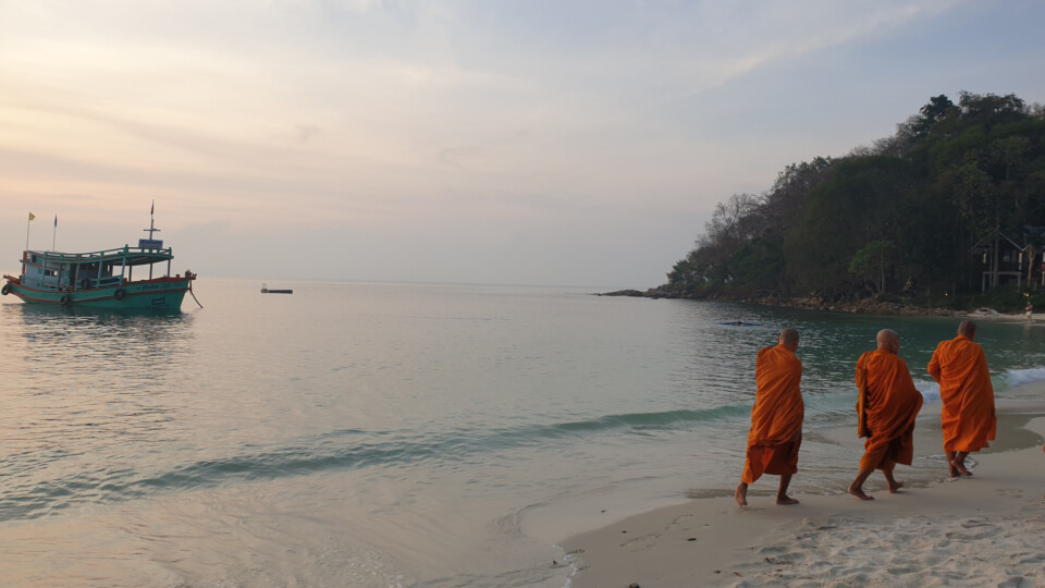 Three monks in orange robes walking along a calm beach at sunrise beside a small anchored boat.