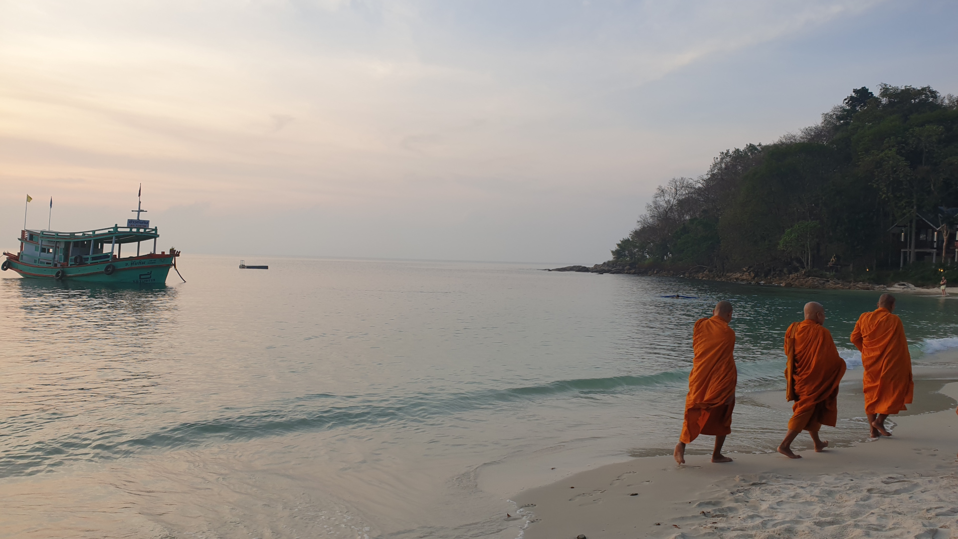 Three monks in orange robes walking along a calm beach at sunrise beside a small anchored boat.