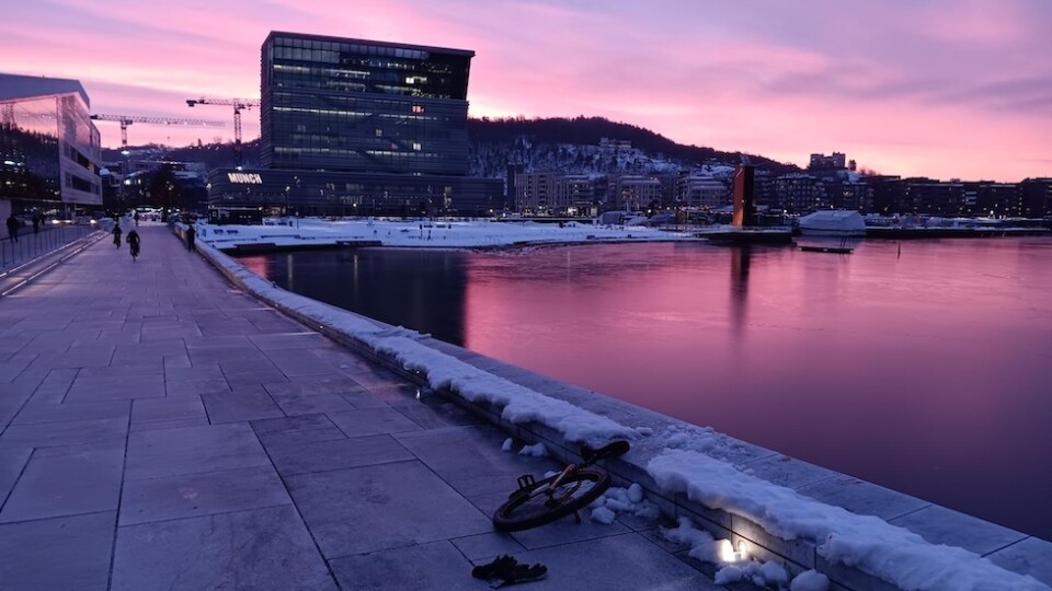 A view looking at the Munch museum. The skies and water (Oslo fjord) are shades of pink and purple in the early morning as the sun begins to rise. There is a unicycle resting on the snowy edge of the path by the water.