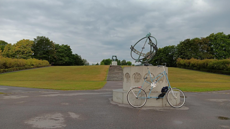 A light blue tallbike leaning against a statue in a park on a cloudy day.