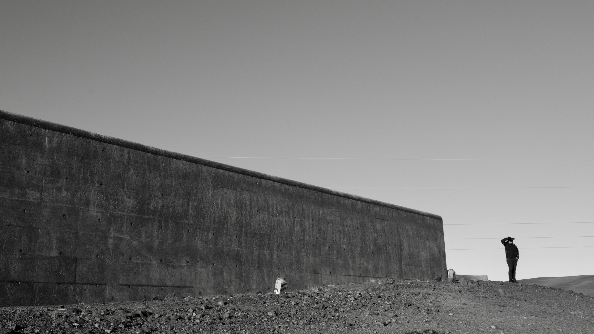 A black-and-white photograph of a massive concrete wall cutting diagonally across a barren, rocky landscape under a clear sky. On the far right, a solitary figure dressed in dark clothing stands on the dirt ground, gazing through binoculars.