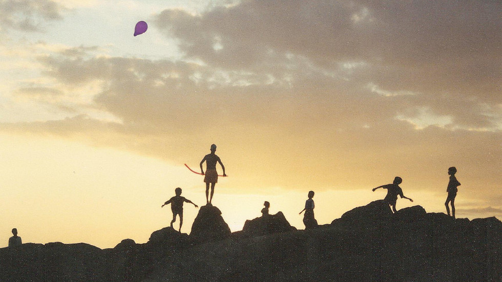 Silhouette of children playing with ballons on rocky ground with a bright sky behind them