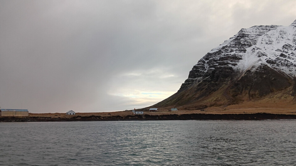 Snow covered hill rising sharply above a rugged shoreline with a few small buildings beside calm water under an overcast sky.