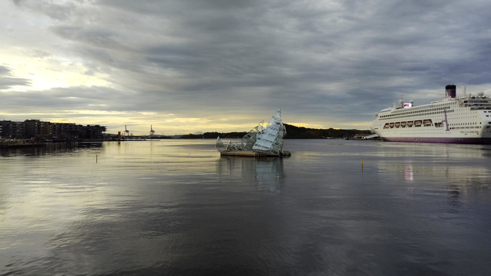 Glass art floats on Oslo fjord, built from sharp, angular forms. A big ship docks to its right, with tall flats on its port. Clouds roll across a moody, bright sky, with light drifting on a calm fjord.