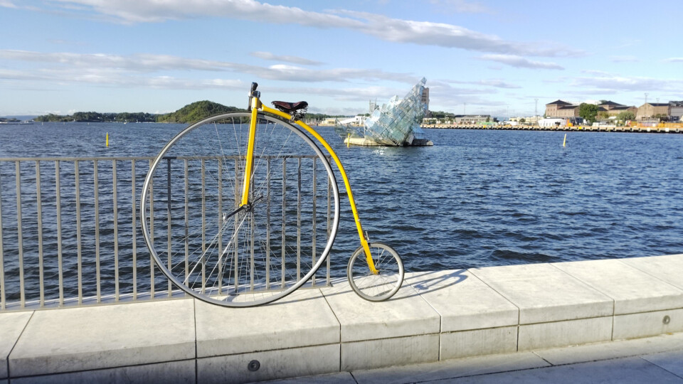 Yellow penny farthing bicycle, leaning on a metal fence beside the waterfront, with the glass sculpture She Lies floating in the Oslo Fjord in the background.