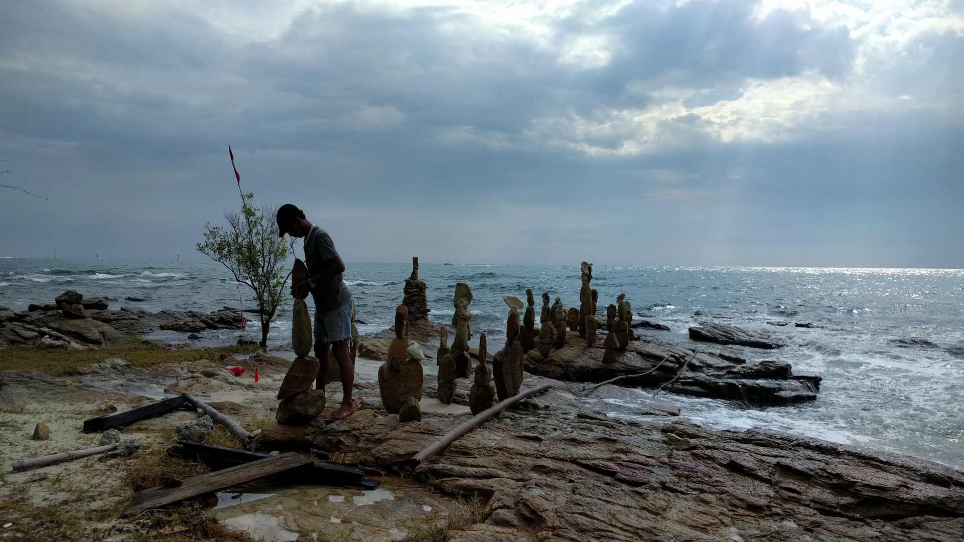 A person balances large stones into vertical sculptures along a rocky shoreline, with the ocean and a partly cloudy sky in the background.