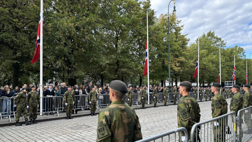 Uniformed Norwegian soldiers stand in formation along Karl Johan as crowds gather behind barricades. Norwegian flags fly at half‑mast during the funeral procession for King Harald V, creating a solemn and respectful atmosphere beneath the tree‑lined street.