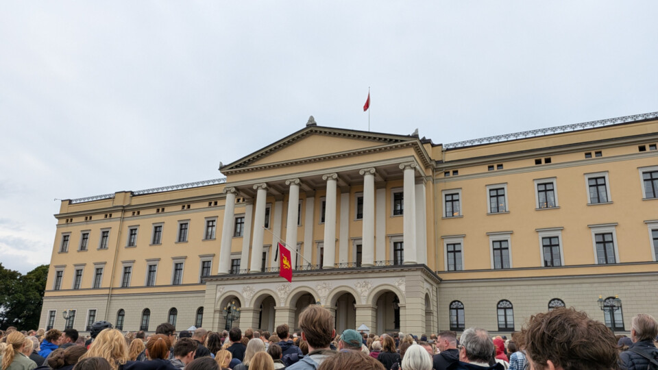 A large crowd stands outside the Royal Palace in Oslo following the death of King Harald. The Norwegian flag flies at full mast while the royal standard is lowered to half‑mast on the palace roof.