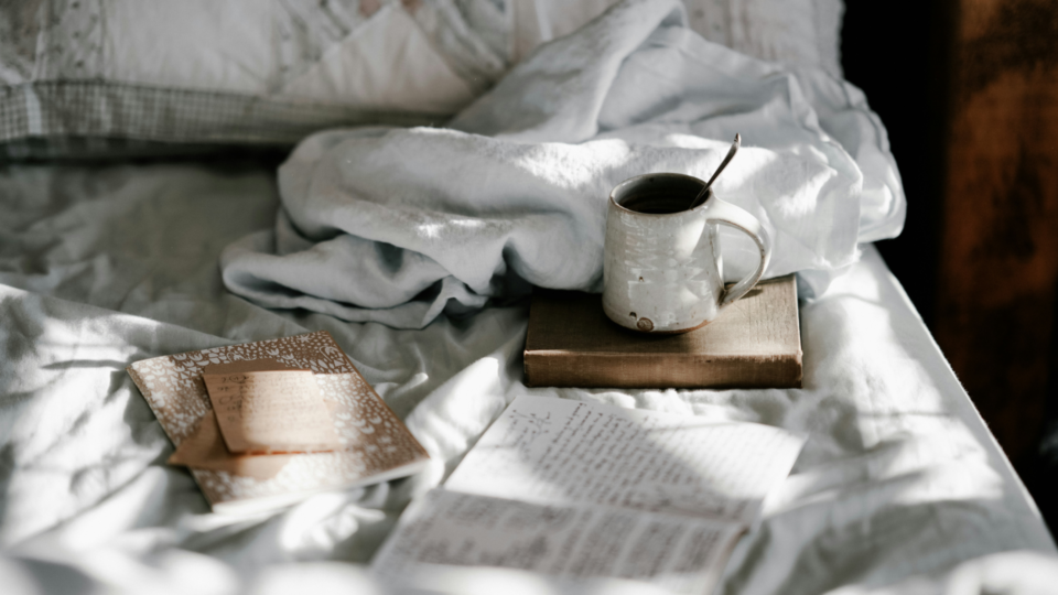 A cozy bed that has some books and notebooks on it. A mug is on one of the books.