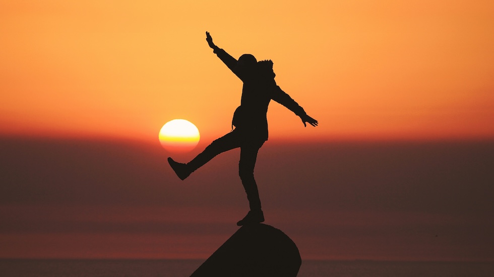 Person in silhouette, balancing on a rock with a sunset inthe background.