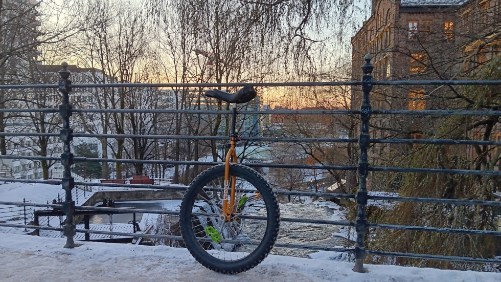 Unicycle resting on the railings of a snow covered bridge. An icy river is visible below. Lots of leafless trees around the edges. Plus some very old industrial buildings on the right bank. In the distance you can see the sun coming up.