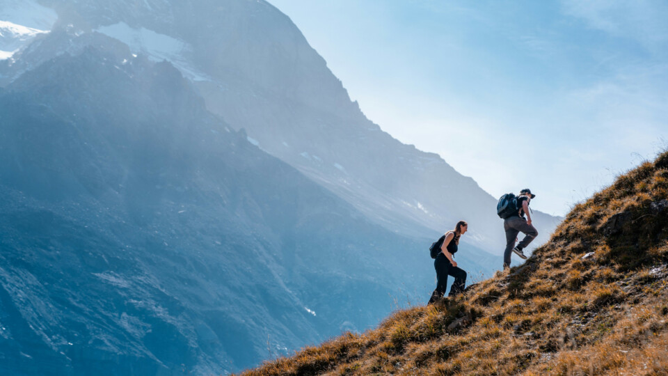 Two people are hiking up a steep grassy hill, with a mountain in the background. Taken near Oeschinen Lake, Kandersteg, Switzerland