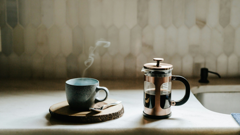 A coffee cup on a coaster and a French press coffee maker on a kitchen counter.