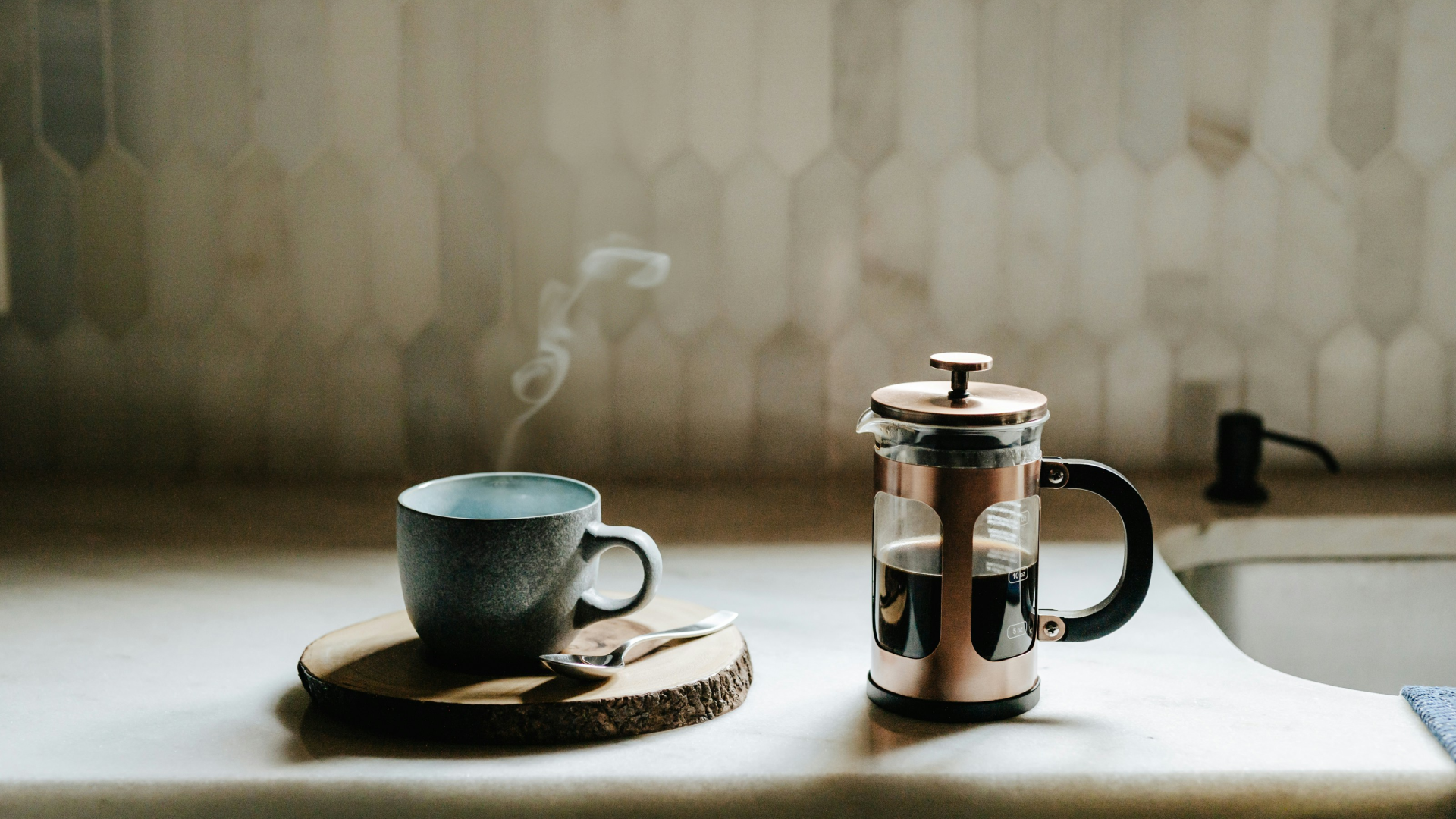 A coffee cup on a coaster and a French press coffee maker on a kitchen counter.