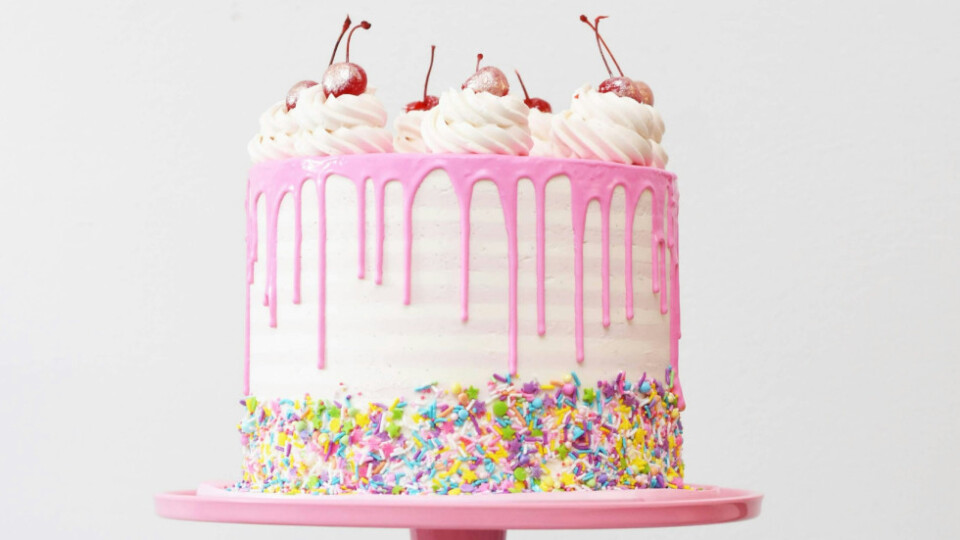 A white cake with pink frosting, and hundreds and thousands around the sides. On top is cream and cherries.