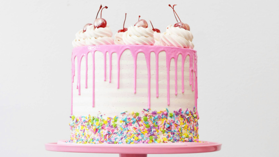 A white cake with pink frosting, and hundreds and thousands around the sides. On top is cream and cherries.