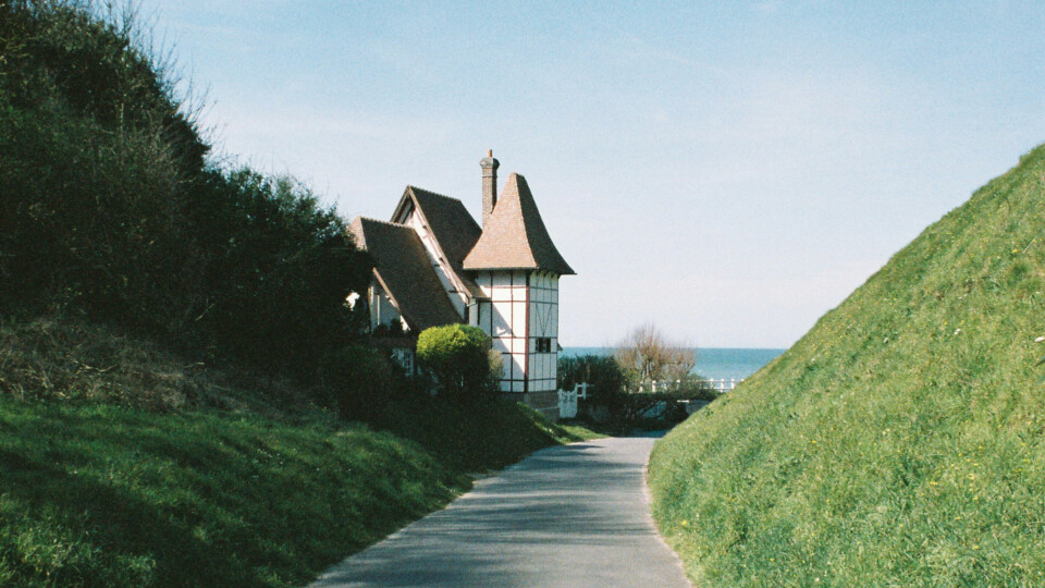 Path to a house between green hills and distant sea.