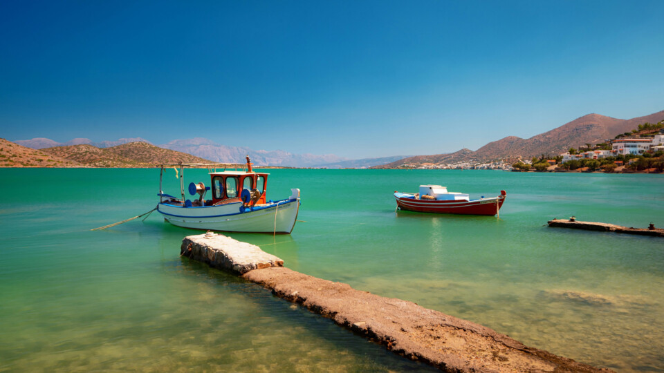 Two small boats floating on calm turquoise water beside a stone pier with hills in the background