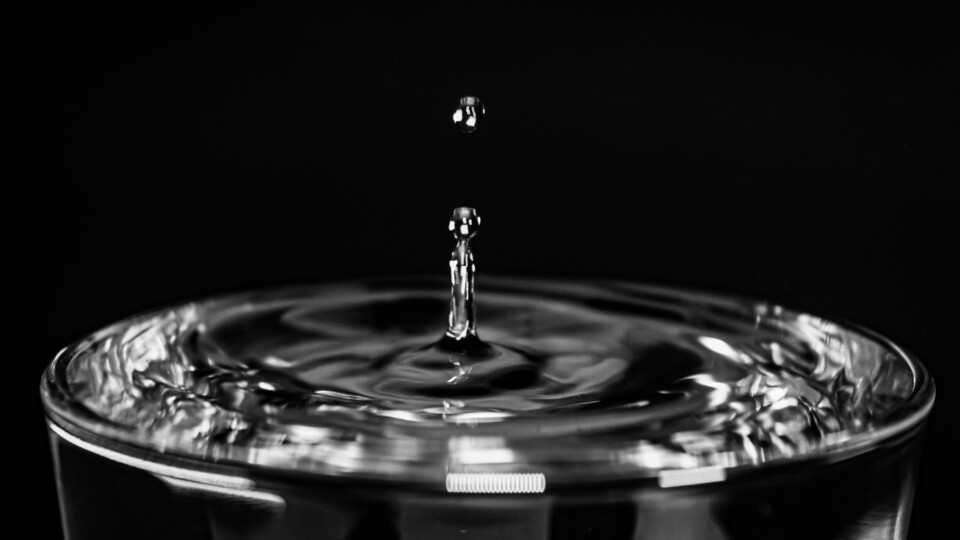 A close‑up shot of a water droplet suspended above a small splash column in a glass against a black background.
