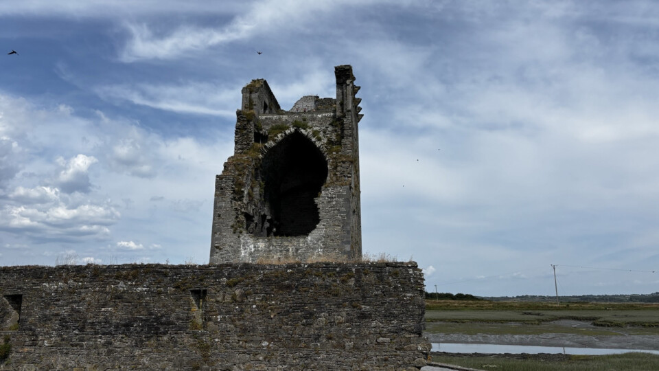 Carrigafoyle Castle on the marshy landscape near Ballylongford, County Kerry. A large arched opening sits high on the weathered stone, with sections of the wall missing. Under a cloudy sky.