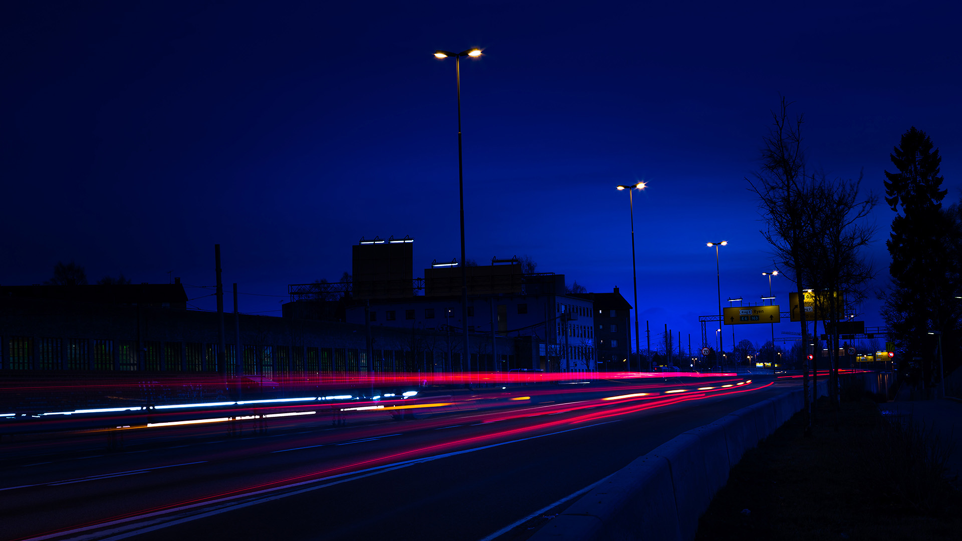 A darkly lit image of a highway. Streaks of head and red tail lights can be seen in the lower part of the frame. In the distance are silhouettes of buildings, road lights and signs.