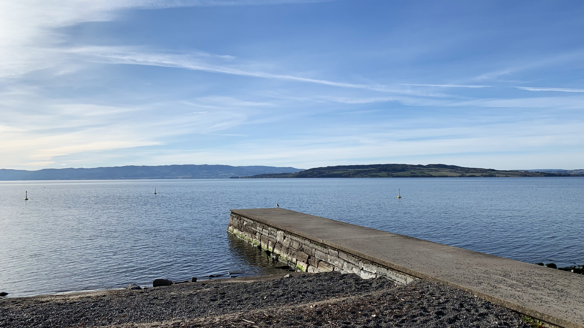 A pier extending from a sandy beach into a large lake. In the distance the other side of the lake can be seen, and in the middle of the lake an island