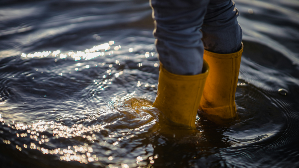 A person wearing blue jeans and yellow wellington boots standing in a deep puddle.