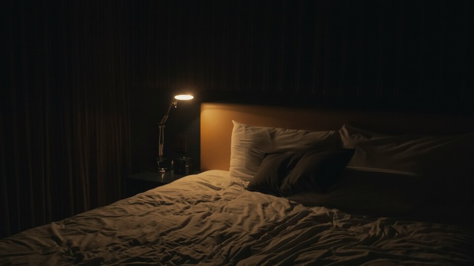 A bed in a dark room with a single side table light on