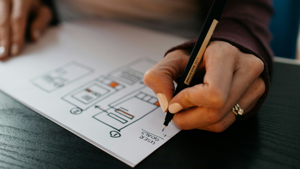 A close-up of a hand holding a black pen, sketching a wireframe layout on paper with orange highlights and writing "User goals" followed by a numbered list.