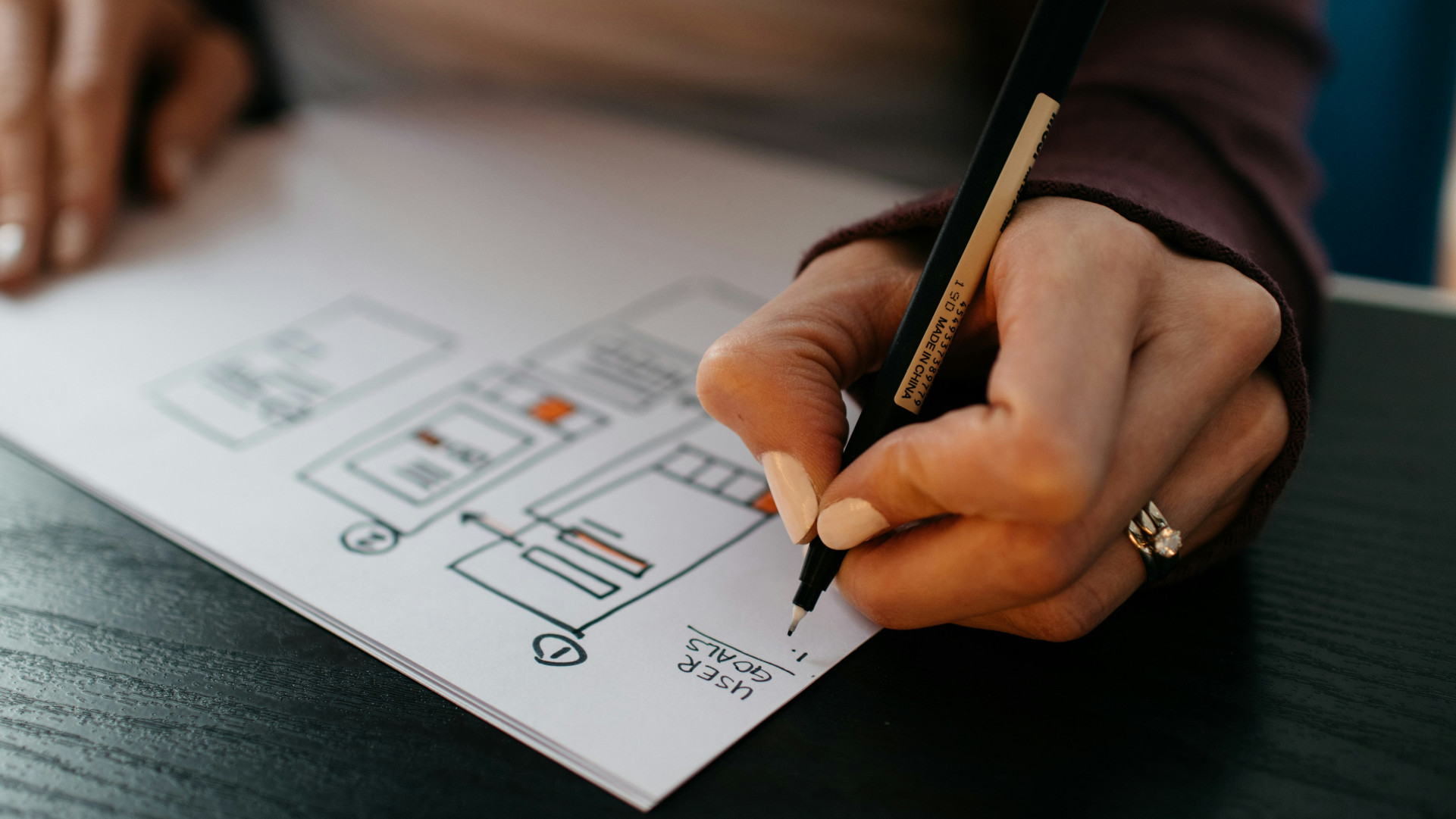 A close-up of a hand holding a black pen, sketching a wireframe layout on paper with orange highlights and writing "User goals" followed by a numbered list.