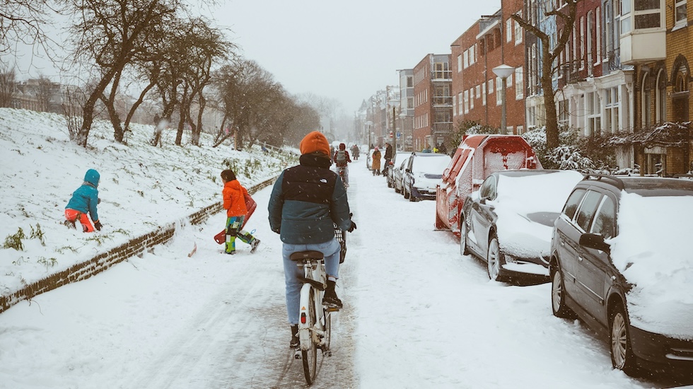 Man cycling along a street in the snow with kids playing at the edge.