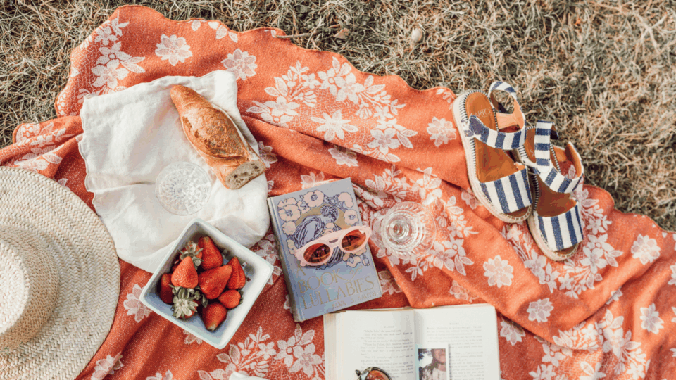 A picnic blanket with various items on it, like a straw hat, sunglasses, books, strawberries, etc.
