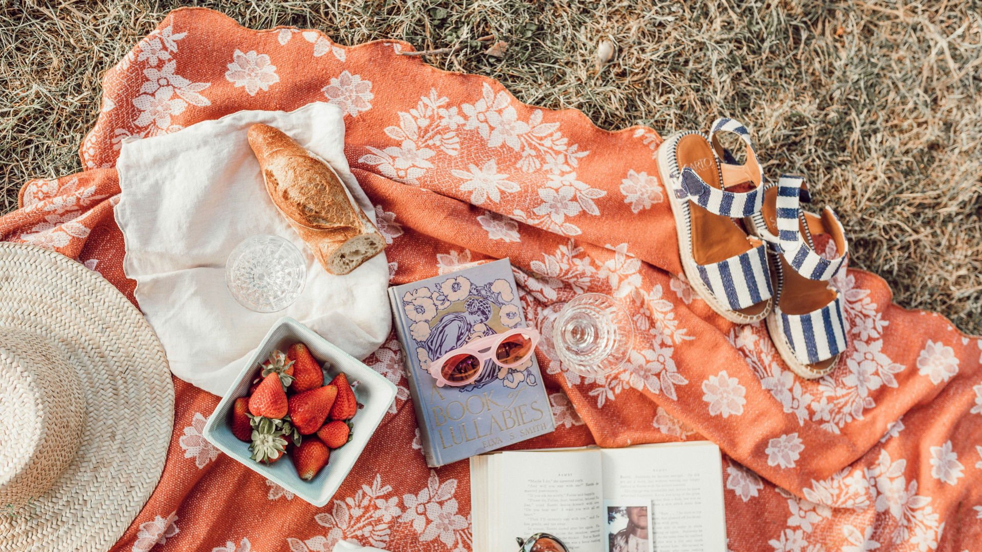 A picnic blanket with various items on it, like a straw hat, sunglasses, books, strawberries, etc.
