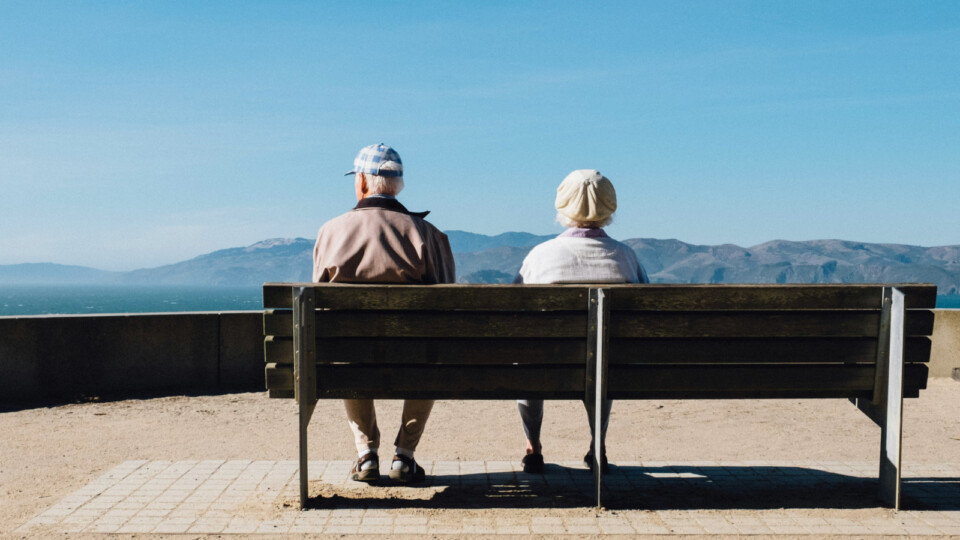 Two older adults sit side by side on a wooden bench overlooking a calm ocean and distant mountains under a clear blue sky, enjoying a peaceful moment outdoors.