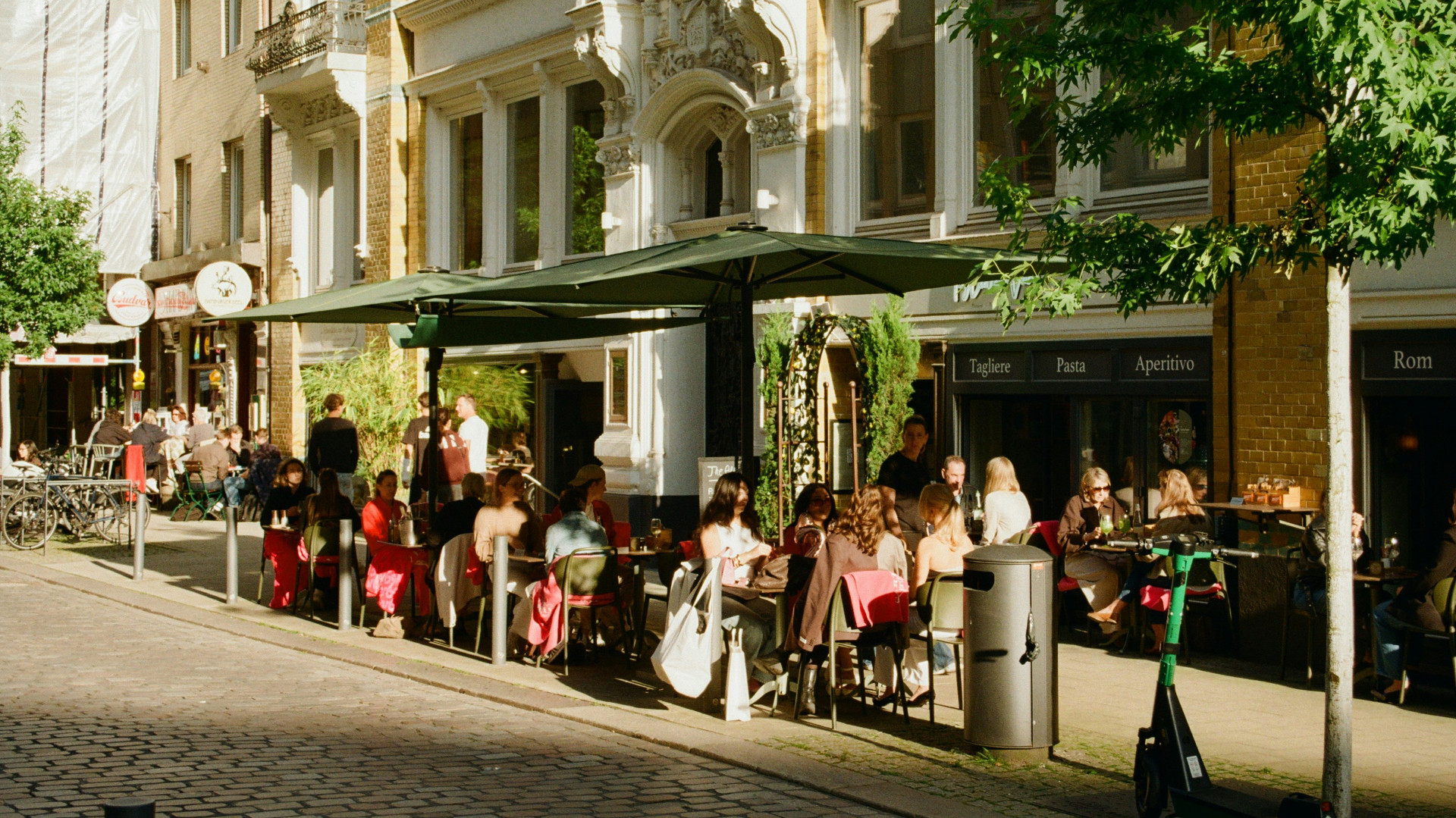 People seated at outdoor café tables beneath large umbrellas along a cobblestone street, with historic buildings, trees, and storefronts illuminated by warm sunlight.