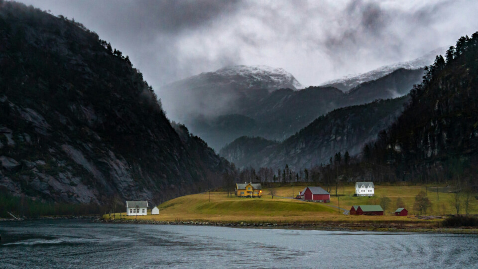Small houses beside a fjord beneath misty mountains and dark cliffs.