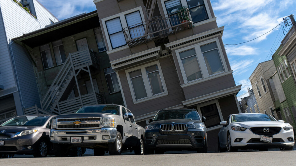 Picture taken from a straight with a sharp include viewed from the side. Parked cars in fron our a house. Due to the rotation of the picture it looks like a flat street with a crooked house.