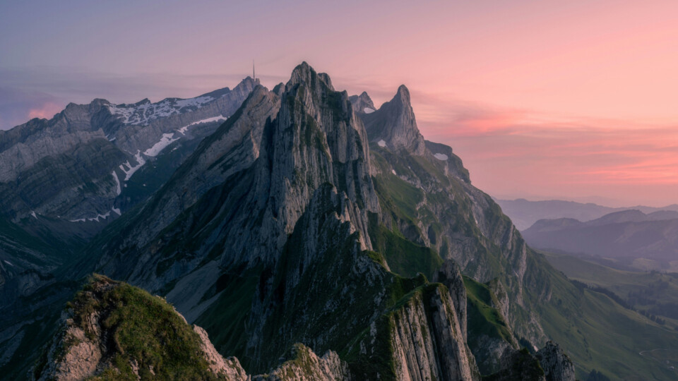 A dramatic view from Schäfler showing jagged alpine peaks glowing in soft pink and purple sunset light, with patches of green and snow across the steep cliffs.