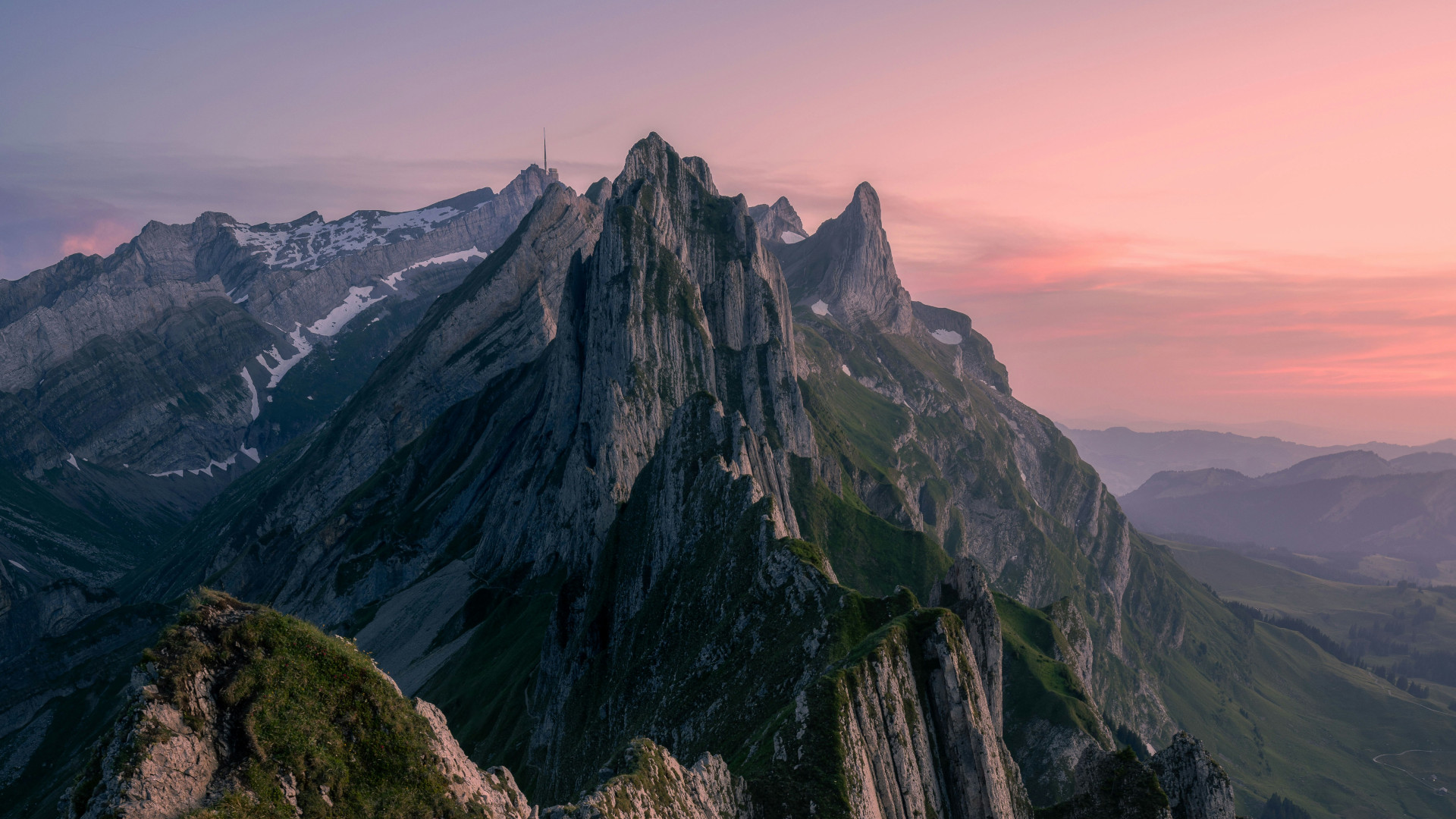 A dramatic view from Schäfler showing jagged alpine peaks glowing in soft pink and purple sunset light, with patches of green and snow across the steep cliffs.