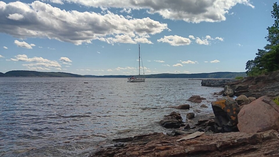 A sailboat on open water on a semi clouded day. There is a rocky coastline in the foreground.