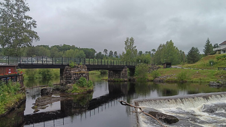 A bridge over a calm river with a small waterfall in the foreground. Cloudy skies.