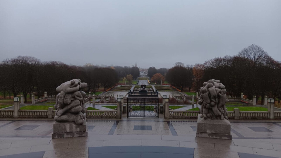 View from the elevated Monolitten plateau in Vigelandsparken, Oslo, looking out over the symmetrical park landscape. In the foreground, two large granite sculptures flank a descending staircase, each depicting intertwined human figures in Gustav Vigeland’s signature style. Beyond the sculptures, formal paths, bare trees, and manicured lawns stretch toward a central fountain, with the overcast sky.