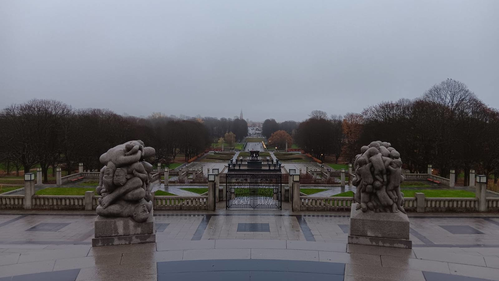 View from the elevated Monolitten plateau in Vigelandsparken, Oslo, looking out over the symmetrical park landscape. In the foreground, two large granite sculptures flank a descending staircase, each depicting intertwined human figures in Gustav Vigeland’s signature style. Beyond the sculptures, formal paths, bare trees, and manicured lawns stretch toward a central fountain, with the overcast sky.