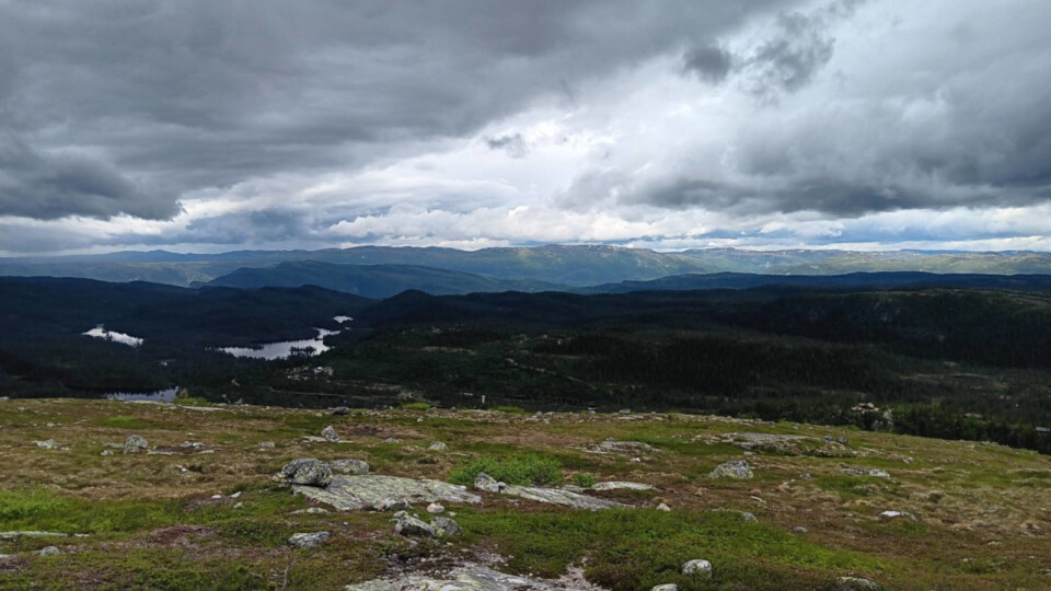 A wide mountain landscape with rocky foreground, dark green forests, small reflective lakes, and distant sunlit ridges beneath dramatic clouds.