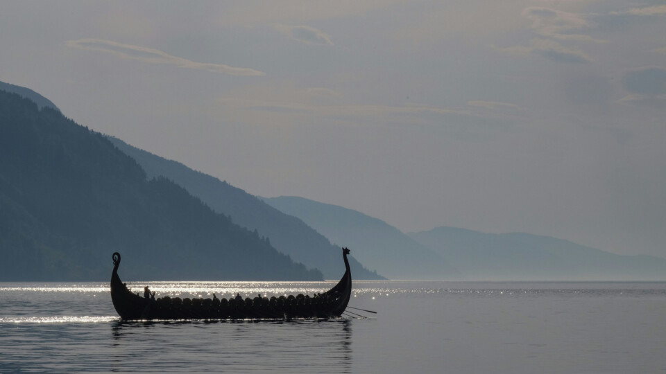 A silhouetted Viking longship gliding across calm water with misty mountains behind it.