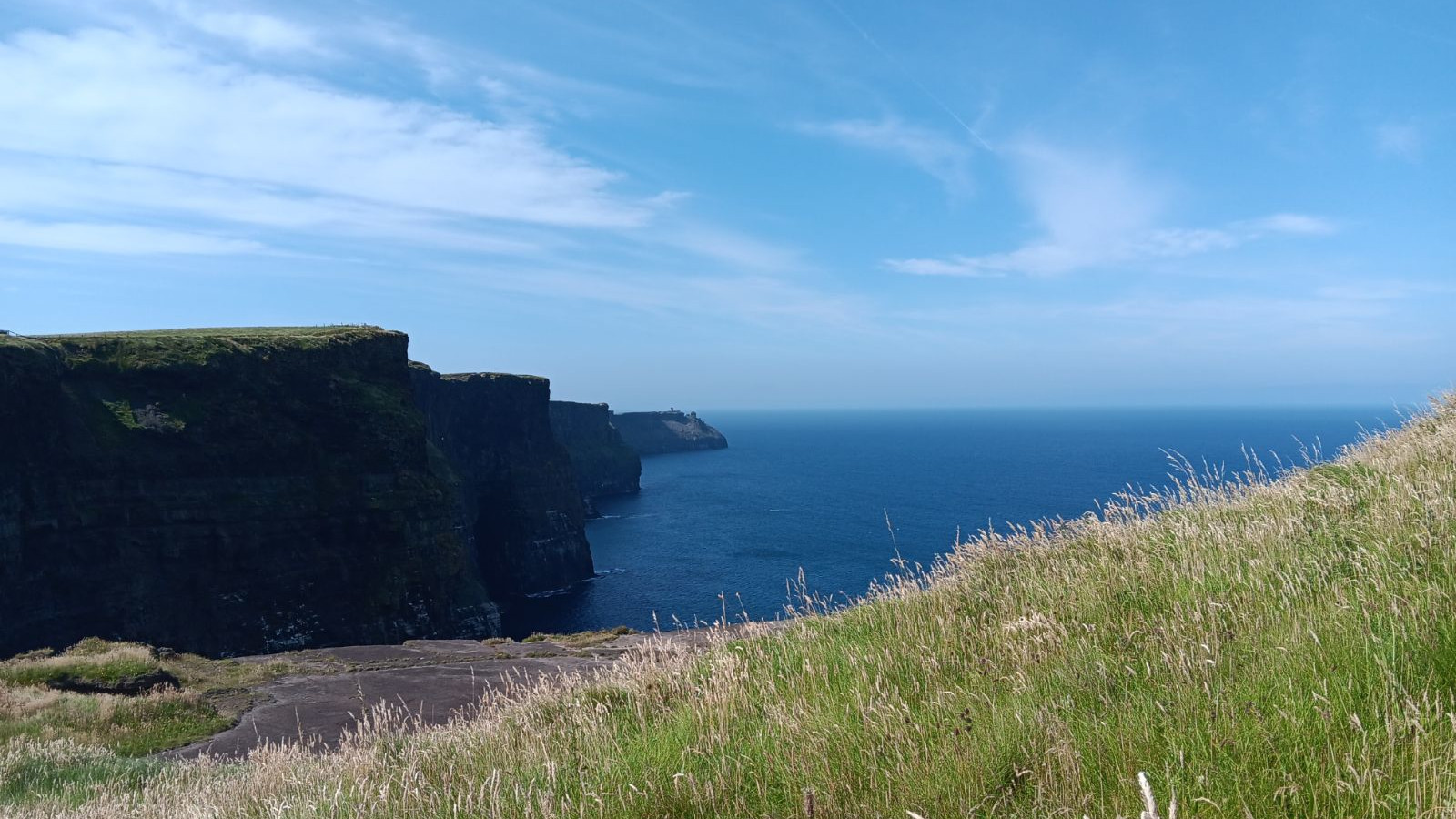 The Cliffs of Moher rising sharply above the Atlantic Ocean, with green grass on the cliff tops and clear blue sky overhead.